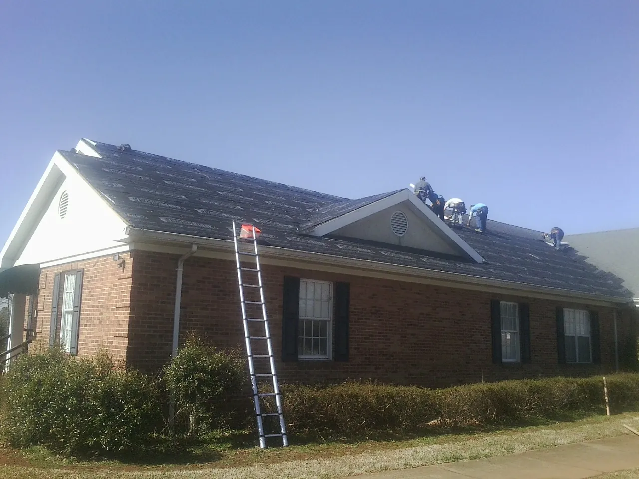 Crew tearing shingles off a brick ranch roof with a ladder set — Emergency Roof Repair work by our crew in East Hampton