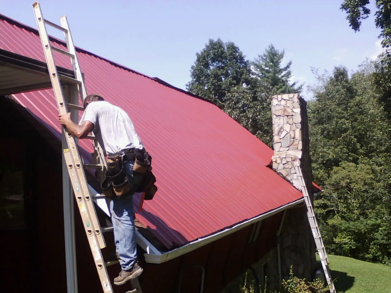 Roofer on a ladder beside a red metal roof and stone chimney — Soffit & Fascia Repair by Roofing Brothers in East Hampton